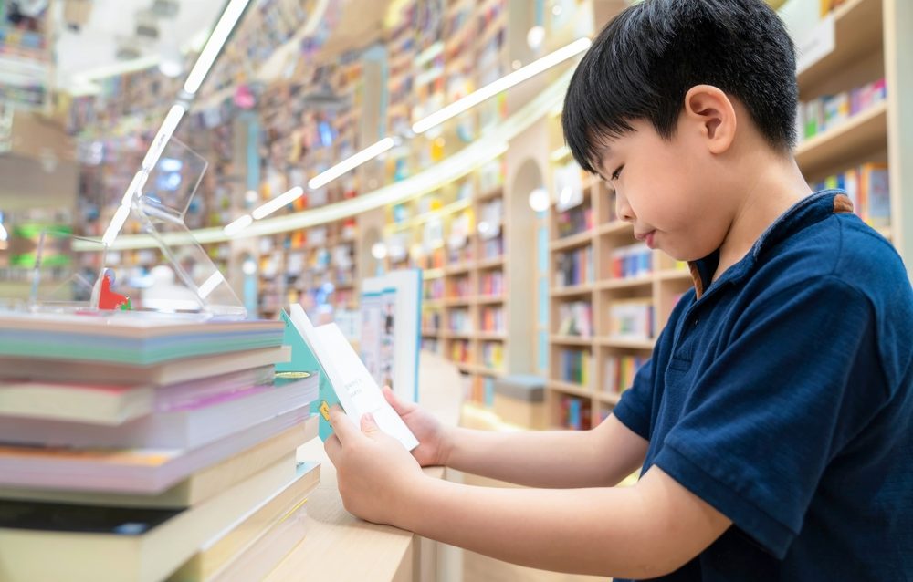 Smart Asia boy sitting on a chair reading a book at multi color bookshelf in the modern and nice interior library with copy space. Many bookshelf at the background. Education, knowledge.