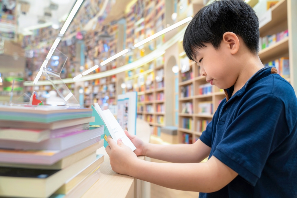 Smart Asia boy sitting on a chair reading a book at multi color bookshelf in the modern and nice interior library with copy space. Many bookshelf at the background. Education, knowledge.